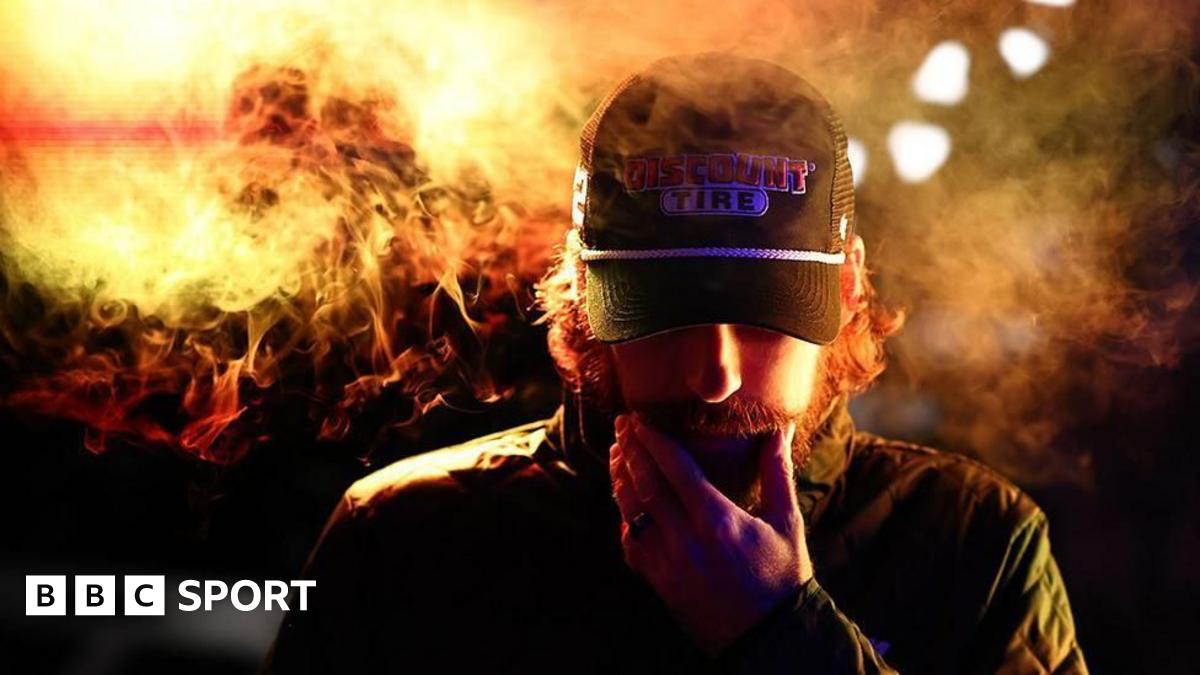 A rugby league player in a green and gold uniform leaps dramatically forward while holding the ball in one hand, scoring a try. Another teammate runs behind, the referee in  a red jersey is nearby. The background shows a packed stadium with spectators.