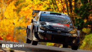 The black Toyota GR Rally1 car of Elfyn Evans above the road surface with a backdrop of golden-leaved trees at Rally Japan