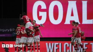 Wrexham players celebrate in a huddle in front of a large scoreboard with the words